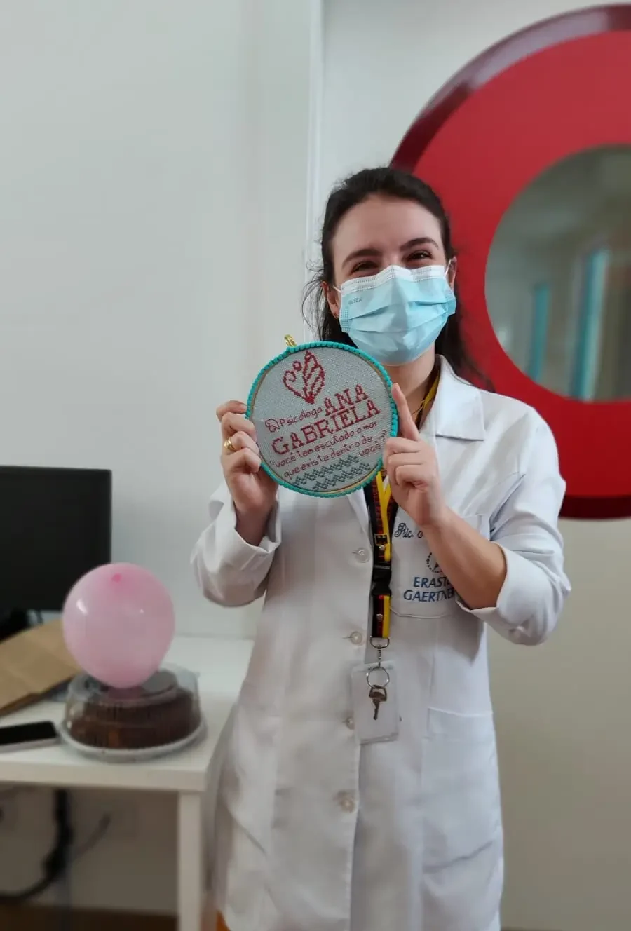 Ana Gabriela Bellaver vestindo jaleco no Hospital Erasto Gaertner, segurando bordado com seu nome e a frase “Você tem escutado o mar que existe dentro de você?”, simbolizando sua escuta sensível, atuação hospitalar e conexão com o cuidado humano.