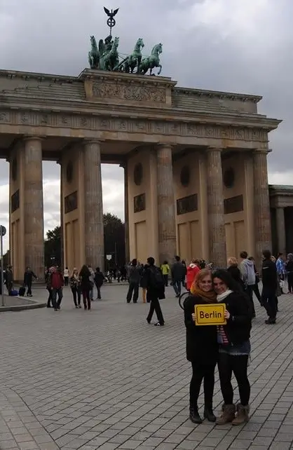 Foto em frente ao Portão de Brandemburgo em Berlim, com duas jovens segurando placa “Berlin”, simbolizando vivência intercultural e a trajetória de Ana Gabriela Bellaver como expatriada.